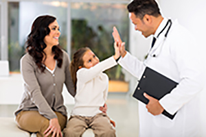 A little girl is giving a male Physician a high five while sitting next to her mom in a doctors office